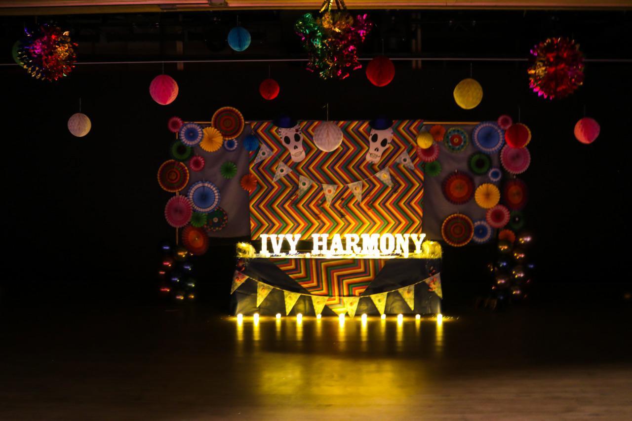 The image showcases a festively decorated stage or event area at IVY STEM International School. Colorful paper decorations like fans, lanterns, and pinwheels create a vibrant and celebratory atmosphere. The illuminated banner with the text 'IVY HARMONY' suggests the name of an event, group, or theme. Candles or small lights along the front edge provide a warm glow. This carefully decorated space is perfect for parties, celebrations, or performances organized by IVY STEM International School.